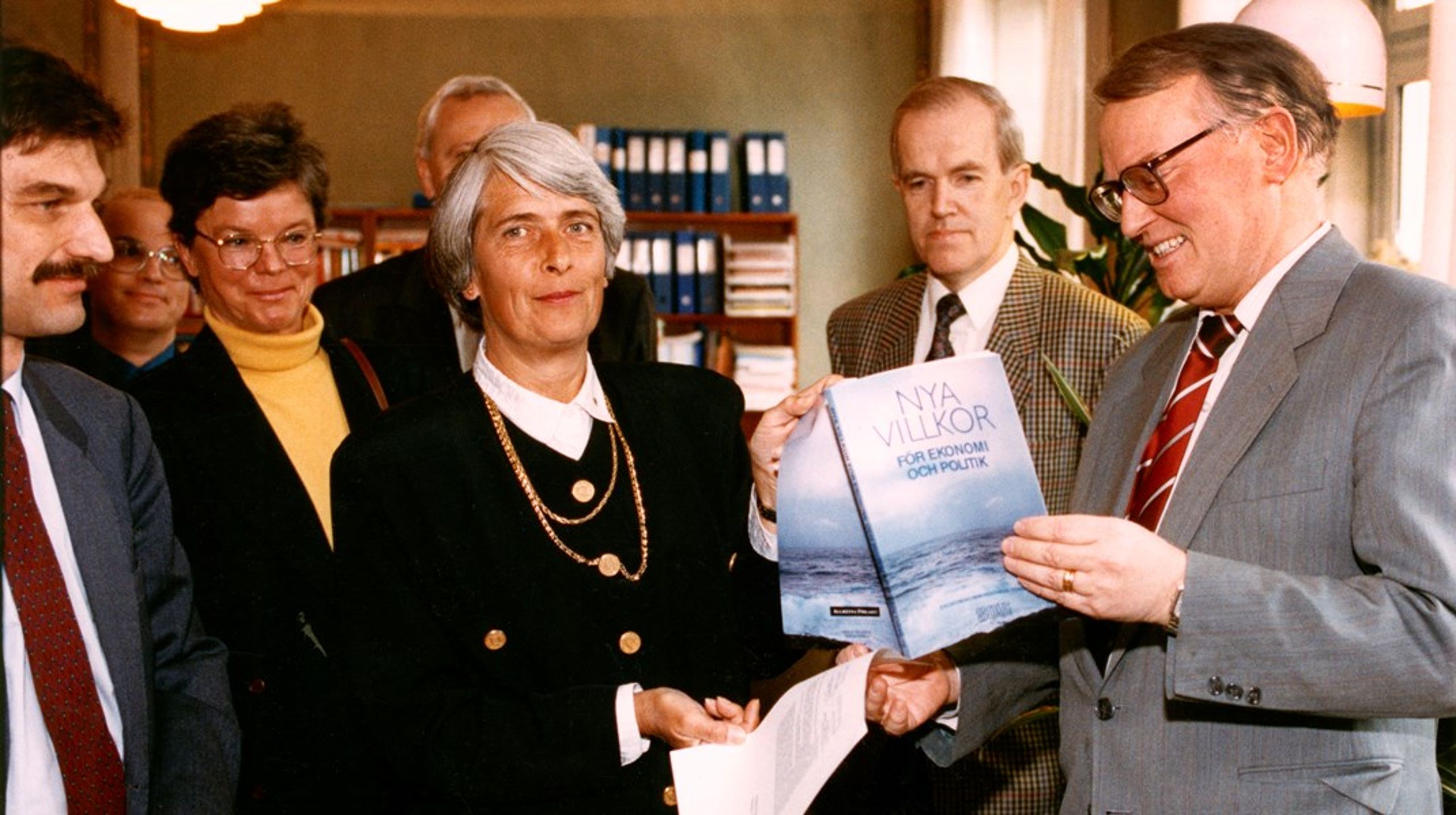 Assar Lindbeck lämnar över utredningen till finansminister Anne Wibble. I vänster bildkant syns Torsten Persson och bakom&nbsp;honom Olof Petersson. Birgitta Swedenborg står till vänster om Wibble.